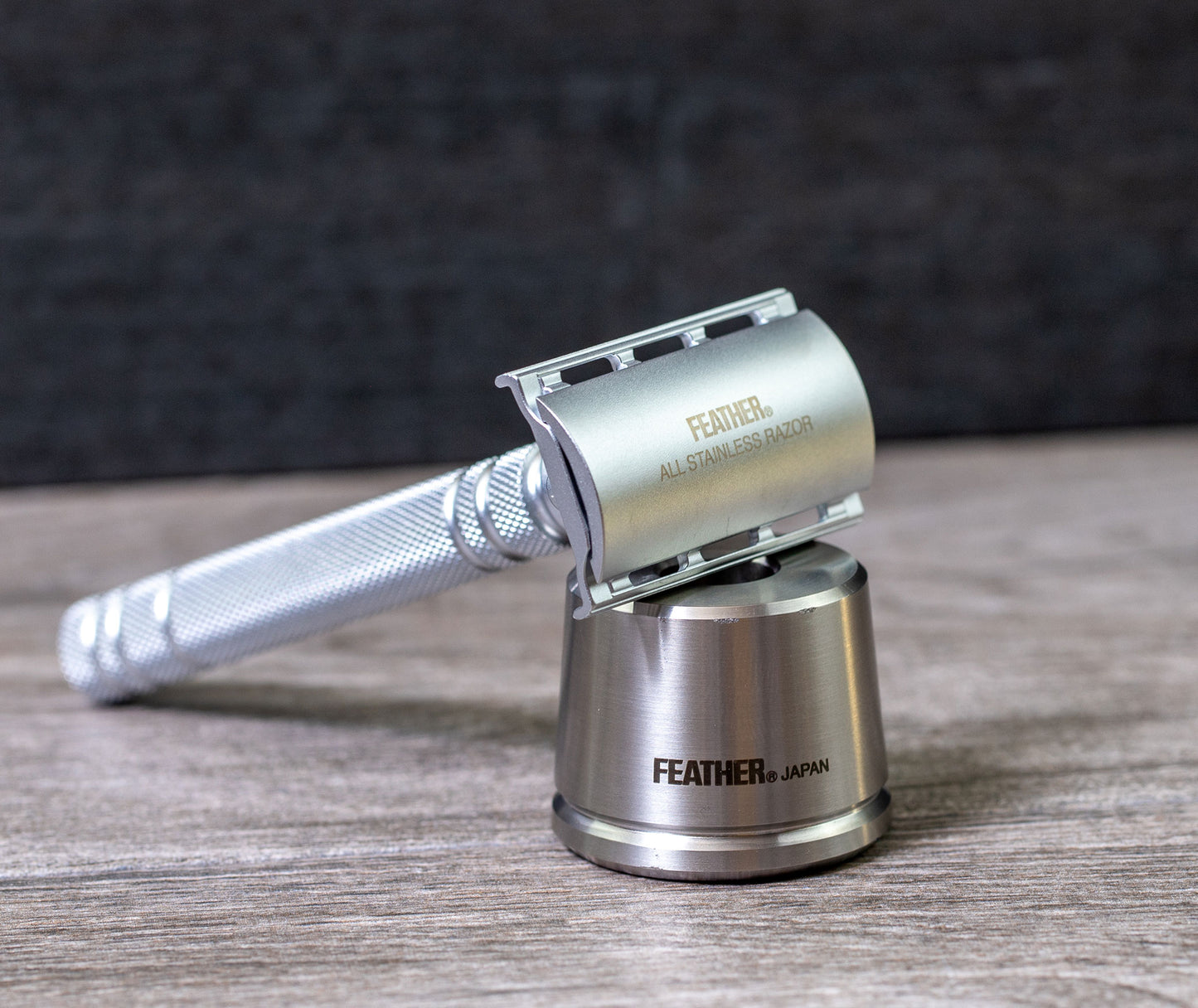 Feather All Stainless Safety Razor placed on its stainless steel stand with a close-up view of the engraved logo.